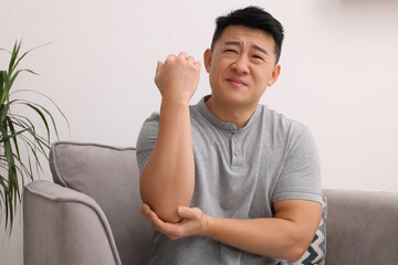 Canvas Print - Asian man suffering from pain in his elbow on armchair indoors