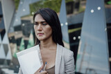 Fototapeta Sypialnia - Thai woman with concerned look holding documents outside office building. Portrait of worried transgender person. Sex discrimination at work, diversity and social inclusion concepts