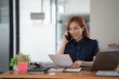 © Songsak C - Young beautiful Asian woman using a smartphone and working with a laptop while sitting at an office desk, working from home concept.