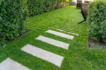 Green lawn and road surface from paving slabs in backyard. Gray color stepping tiles floor, strip line concrete pavement and green grass carpet lawn. Concrete paving with tiles and cut grass in park.