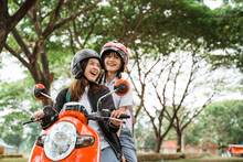 Two Student Girls Wearing Helmets And Jackets Chatting While Riding Motorbikes Together