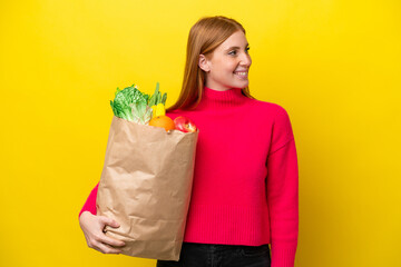 Wall Mural - Young redhead woman holding a grocery shopping bag isolated on yellow background looking side