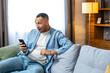 © Ivan - Surprised happy african man holding phone looking at cellphone read good news in sms sit on sofa, amazed black guy winner excited by mobile app win scream with joy celebrate victory success at home