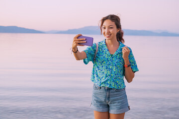 Wall Mural - Happy girl travel blogger taking selfie photo on a high cliff with splendid view of panoramic seascape or lakeshore and mountains.