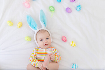 Wall Mural - happy baby boy with rabbit ears on his head lying on the bed with Easter eggs, cute funny smiling little baby. The concept of Easter. Space for text