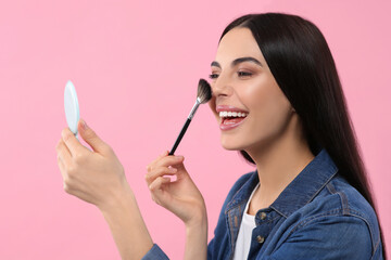 Wall Mural - Beautiful woman with cosmetic pocket mirror applying makeup on pink background