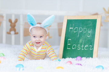 Wall Mural - baby boy with rabbit ears on his head lies with the inscription Happy Easter on the bed with Easter eggs, cute funny smiling little baby. The concept of Easter.