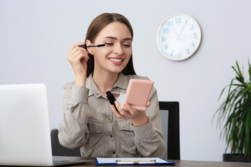 Sticker - Young woman with cosmetic pocket mirror applying mascara at table indoors