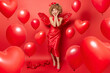 Leinwanddruck Bild - Horizontal shot of beautiful romantic woman keeps lips folded expresses love to you wears dress and wings prepares for St Valentines Day surrounded by inflated balloons isolated over red background