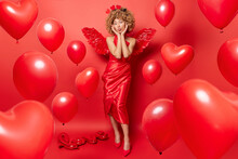 Horizontal Shot Of Beautiful Romantic Woman Keeps Lips Folded Expresses Love To You Wears Dress And Wings Prepares For St Valentines Day Surrounded By Inflated Balloons Isolated Over Red Background