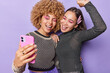 © wayhome.studio  - Two positive young women have fun and pose for making selfie wear bright makeup fashionable clothes laugh joyfully get prepared for party isolated over purple background. Friendship concept.