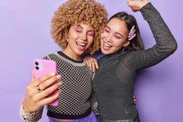 Wall Mural - Two positive young women have fun and pose for making selfie wear bright makeup fashionable clothes laugh joyfully get prepared for party isolated over purple background. Friendship concept.