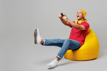 Wall Mural - Full body young man of African American ethnicity he wear pink t-shirt yellow hat headphones sit in bag chair hold in hand play pc game with joystick console isolated on plain grey background studio.