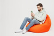 © ViDi Studio - Full body young satisfied happy fun cheerful man wear mint hoody sit in bag chair hold in hand use mobile cell phone isolated on plain solid white background studio portrait. People lifestyle concept.