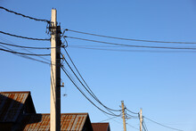 Powerline Posts With Electrical Wires And Capacitors Above Old Roofs On Blue Sky Background. Electricity Transmission Line, Power Supply In Village