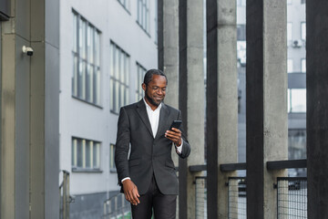 Happy successful African American businessman boss walking outside office building, manager holding smartphone reading message and smiling, using communication app.