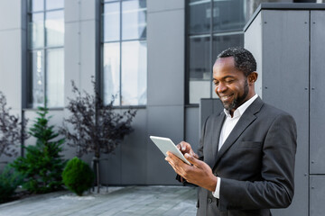 Successful african american man in business suit smiling and using tablet computer, businessman outside modern office building walking.