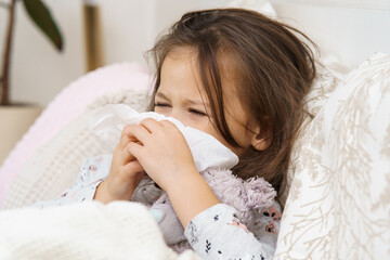 Little girl with closed eyes blow stuffy nose with handkerchief. Portrait of unhealthy child. Kid in sleepwear lie under blanket and have bed rest. Home treatment. Runny nose, snot, cold symptoms.