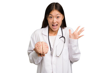 Wall Mural - Young asian doctor woman holding a brain isolated receiving a pleasant surprise, excited and raising hands.