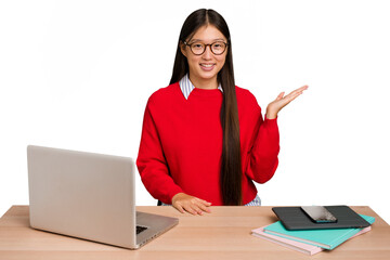 Wall Mural - Young student asian woman in a workplace with a laptop isolated showing a copy space on a palm and holding another hand on waist.