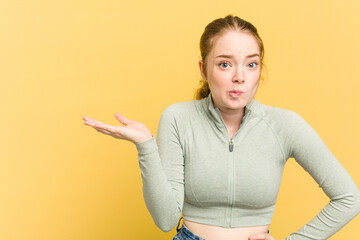 Wall Mural - Young caucasian redhead woman isolated on yellow background showing a copy space on a palm and holding another hand on waist.