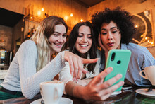 Group Of Young Women Having Fun Sharing Media With An Cellphone. Three Girls Looking To The Smartphone On A Coffee Shop, Restaurant Or Bar. Female Students Buying On A Market Place Using A Mobile