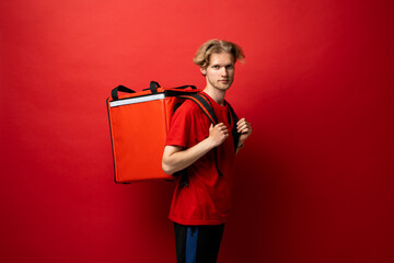 Wall Mural - Best Delivery Service Concept. Portrait of confident smiling male courier wearing red uniform and thermo backpack bag looking at camera isolated on red background.