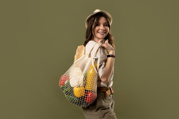 Wall Mural - Smiling young woman in light summer clothes with a mesh eco bag full of vegetables, greens on a green studio background. Sustainable lifestyle. Eco friendly concept. Zero waste.