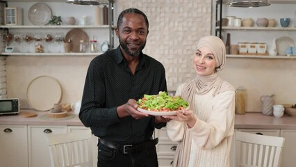 Wall Mural - Attractive african american man and muslim young woman holding bowl with healthy salad with fresh vegetables while cooking dinner together and having fun in a new light kitchen.