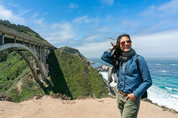 Canvas Print - stylish smiling asian Taiwanese lady tourist holding blowing hair while enjoying comfortable sea wind and scenic view near big sur bridge in California usa