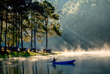 Man Sail The Small Blue Boat In The Lake Pass Through Beam Light That Shine Through Tree In Forest Of National Park In Early Morning With Mist Cover The Water And Look Beautiful For Travel And Relax.