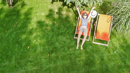 Woman relaxes in summer garden in sunbed deckchair on grass, young girl outdoors in green park on weekend, aerial drone view from above
