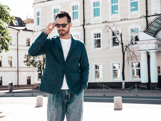 Wall Mural - Handsome confident stylish hipster lambersexual model. Sexy modern man dressed in elegant blue suit and jeans. Fashion male posing in the street background in Europe city at sunset. In sunglasses