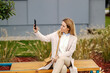 © Dusan Petkovic - A cute blond fashionable woman is sitting on a bench in park and taking selfies.