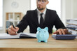 © Studio Romantic - Piggy bank on desk of accountant who uses calculator to analyze budget and make calculations. Close up of blue piggy bank in form of pig on background of man making entries in accounting documents.