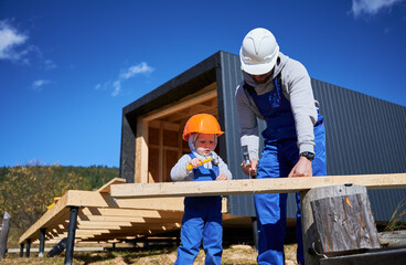 Wall Mural - Father with toddler son building wooden frame house. Male builders hammering nail into plank on construction site, wearing helmet and blue overalls on sunny day. Carpentry and family concept.