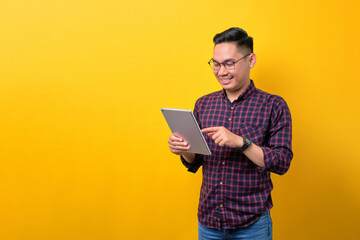 Smiling young Asian man in glasses using digital tablet, browsing internet on modern gadget isolated over yellow background