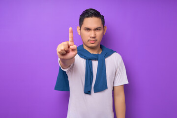 Wall Mural - angry young Asian man wearing white t-shirt looking at camera and showing stop gesture with his index finger isolated over purple background. people lifestyle concept