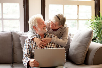 Wall Mural - Lovely senior couple relax on sofa in living room enjoying retirement and free time browsing on laptop. Older generation and wireless technology users concept