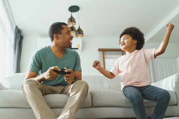 Wall Mural - Cheerful Black African American Father and his Little son in Afro hair sitting on sofa using joysticks or game controllers playing console video games