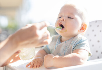 mother spoon feeding her baby boy child in baby chair with fruit puree on a porch on summer vacation