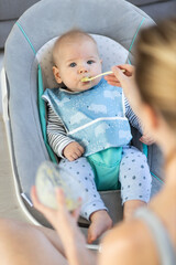 Wall Mural - Mother spoon feeding her baby boy child in baby chair with fruit puree. Baby solid food introduction concept