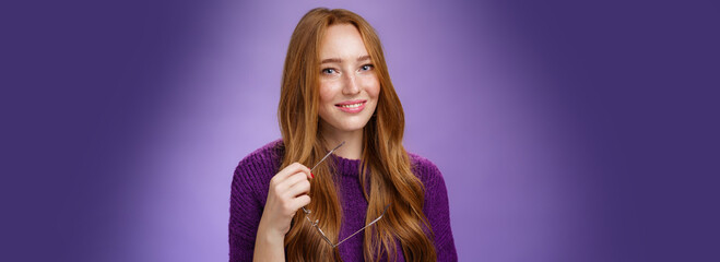 Wall Mural - Close-up shot of charming tender and cute redhead woman in purple sweater holding glasses and smiling broadly as having nice friendly conversation against violet background