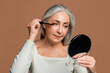 © Prostock-studio - Cheerful senior caucasian female with gray hair applies mascara on eyelashes with brush, looks in mirror