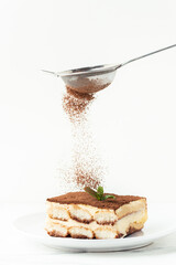 Wall Mural - Pastry Chef sprinkles cocoa powder through a sieve on a tiramisu cake decorated with green mint. Making traditional Italian no-baking dessert. White background