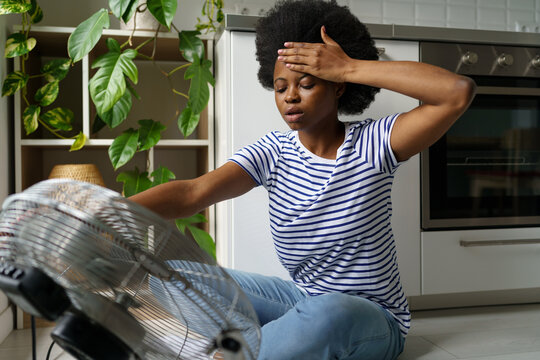 unhappy african woman touching forehead having hot flashes using electric fan to cool off, overheate