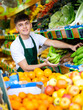Positive young male supermarket worker in green apron arranging bananas in the fruit section