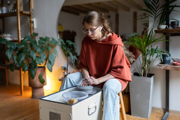 Young woman wearing glasses learning how to throw clay on pottery wheel, visiting ceramics masterclass. Female potter ceramicist at workplace in cozy home-based studio, producing earthenware at home