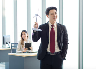 Millennial Asian young professional successful male businessman manager in formal suit standing smiling holding windmill model showing thumb up in company office while colleague working at workplace