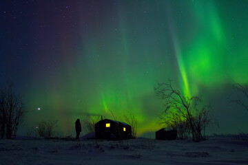 Wall Mural - Winter Arctic landscape with the aurora borealis over the snowy tundra. A man stands near the hunting lodge and admires the northern lights. The planets Venus and Jupiter are visible above the horizon